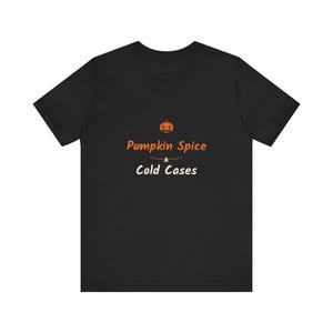 May include: A black short-sleeve t-shirt featuring a graphic centered on the chest. The design includes a small orange pumpkin icon above the orange text Pumpkin Spice, a small horizontal line, and the white text Cold Cases below it.