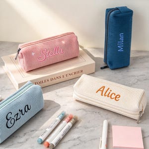 May include: Four rectangular corduroy pencil cases in pink, blue, cream, and dark blue. Each case features a zipper closure and is personalized with embroidered names: Stella, Ezra, Alice, and Milan. The cases are arranged with markers.