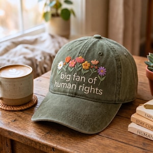 以下が含まれることがあります： オリーブグリーンの野球帽で、カラフルな花の刺繍と白い文字で「big fan of human rights」と書かれています。この帽子は構造化されたクラウン、カーブしたつば、通気のための上部パネルに縫い付けられたアイレットが特徴です。
