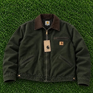 May include: A dark olive green canvas jacket featuring a brown corduroy collar, a full-length front zip, and a chest pocket with a zip closure. A rectangular Carhartt brand patch is visible on the chest, and a paper tag hangs from the zip pull.