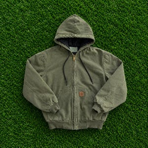 May include: An olive green hooded jacket with a full-length front zip, black drawstring cords, and a quilted black interior lining. The jacket features a front kangaroo pocket with a small rectangular brown brand patch on the lower right side.