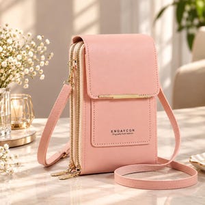 May include: A light pink crossbody phone bag with a fold-over flap, gold-tone hardware, and two zippered compartments. The front features the text ENDAYCON Originally from fashion. It includes a matching adjustable shoulder strap and side rings.