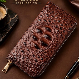 May include: A rectangular brown leather wallet featuring a prominent embossed crocodile skin texture with a central scale pattern. The wallet includes a gold-toned metal zip closure with a matching pull tab and the text PEAK LEATHERS CO.