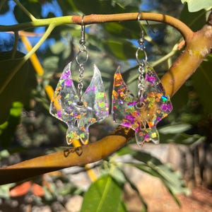 May include: A pair of clear resin bat-shaped earrings featuring embedded iridescent purple, orange, and green foil flakes. The earrings hang from silver-toned metal hooks and short chain links, showcasing a translucent finish with reflective colourful accents.