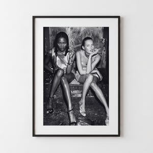 Könnte beinhalten: Ein gerahmtes Schwarz-Weiß-Foto, das zwei Personen zeigt, die nebeneinander sitzen. Beide tragen ärmellose Oberteile und helle Schuhe. Eine Person hat langes dunkles Haar und Schmuck, die andere helles Haar, beide halten Zigaretten in den Händen.