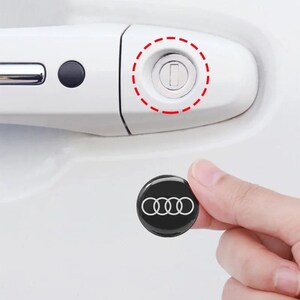 May include: A small, round, black adhesive cover featuring the white Audi four-ring logo. The emblem is designed to be placed over the circular keyhole on a white car door handle, which is highlighted by a red dashed circle.