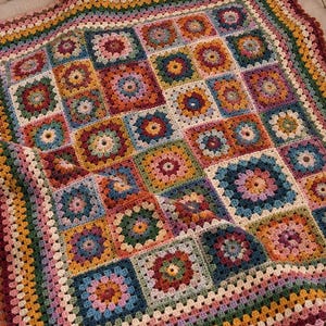 Może przedstawiać: Ręcznie robiony szydełkowy koc z kolorowym wzorem typu granny square. Kwadratowe motywy są ułożone w siatkę, prezentując różnorodne kolory włóczki, w tym musztardowy żółty, ciemnoczerwony, leśny zielony, granatowy oraz delikatne odcienie różu.