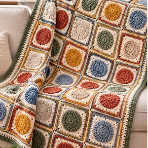 May include: A crochet blanket featuring a repeating pattern of square motifs, each containing a circular center in shades of rust, mustard yellow, sage green, and dusty blue, framed by cream-colored yarn and a dark green border.