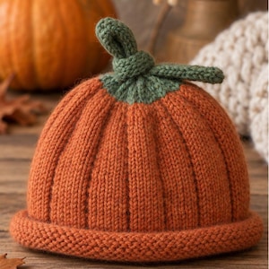 Puede incluir: Un gorro de punto naranja con textura acanalada y borde enrollado, que presenta un detalle de tallo de punto verde en la parte superior. El gorro está diseñado para parecerse a una calabaza, con nervaduras verticales desde la base al tallo.