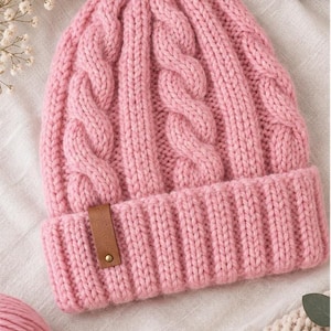 May include: A soft pink knit beanie featuring a vertical cable knit pattern and a wide ribbed cuff. A small rectangular brown faux leather tag with a single metallic rivet is attached to the front of the cuff, adding a subtle contrast to the yarn.