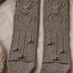 Könnte beinhalten: Ein Paar taupefarbene Strickstrümpfe mit einem strukturierten Zopfstrick-Spinnenmuster im oberen Bereich und drei kleineren, erhabenen Spinnenmotiven, die an vertikalen Linien im unteren Teil hängen, abgeschlossen mit einem gerippten Bündchen.