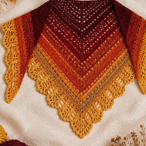 Könnte beinhalten: Ein dreieckiges Häkeltuch mit einem Farbverlaufsmuster, das von tiefem Burgunderrot in der Mitte zu leuchtendem Orange und Goldgelb an den Rändern übergeht, abgeschlossen mit einer dekorativen, bogenförmigen Spitzenkante am unteren Rand.