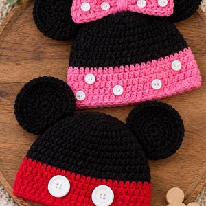 May include: Two crocheted hats with round black ears. The top hat is black with a pink brim and a pink bow, both featuring white buttons. The bottom hat is black with a red brim and two white buttons. A small wooden mouse-shaped cutout is included.