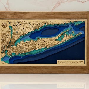 May include: A framed 3D wooden bathymetric map of Long Island, NY, featuring layered blue water depths and a detailed land topography with engraved city names, road networks, and a compass rose, all encased in a natural light oak wood frame.