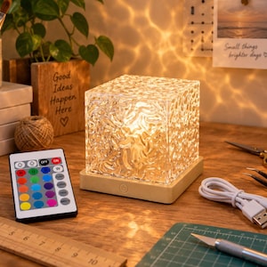 May include: A square, textured glass lamp sits on a light-colored wooden base with a power button. It includes a remote control with multiple color buttons, a white USB charging cable, and a wooden ruler measuring up to 10 inches in length.