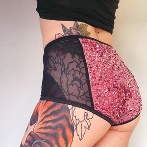 Puede incluir: Ropa interior de cintura alta rosa y negra con un panel de lentejuelas y un panel de malla transparente. La ropa interior tiene una cintura el&aacute;stica negra.