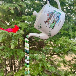 May include: A white ceramic teapot bird feeder with a gold rim, featuring a colorful illustration and the word "GALWAY". The teapot is decorated with shamrocks and suspended by a black cord. A beaded strand with green, white, and gold beads hangs from the spout.