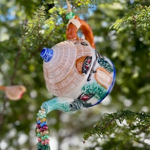 May include: A ceramic teapot shaped like a house with a colorful painted design hangs from a tree branch. The teapot has a beaded chain hanging from the spout.