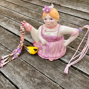 May include: A whimsical ceramic teapot in the shape of a woman wearing a pink dress and a floral crown. The teapot has a yellow spout and is holding a beaded necklace and a pink cord. The teapot is on a wooden surface.