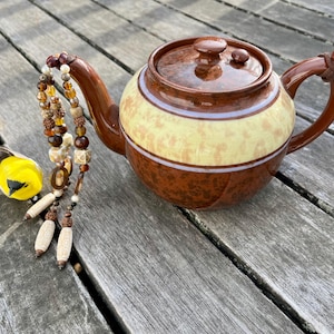 May include: A brown teapot with a light brown and white patterned design sits on a wooden surface. A beaded necklace with brown, white, and orange beads hangs next to the teapot.