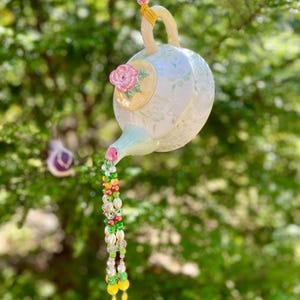 May include: A hanging, decorative teapot ornament with a floral design and a pink rose accent. The teapot is cream-colored with a yellow handle and spout, and a beaded stream of colorful beads flows from the spout.