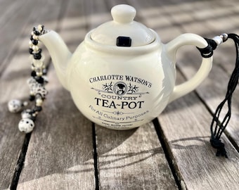 May include: White ceramic teapot with black lettering that reads "Charlotte Watson's Country Tea-Pot For All Culinary Purposes". The teapot has a black string attached to the handle.