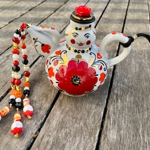 May include: A white ceramic teapot with a red and orange floral design, a black tassel, and a red flower on the lid. The teapot has a painted face with black eyes and a red mouth.
