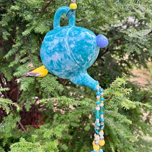 May include: A turquoise and white marbled ceramic bird feeder shaped like a teapot, hanging from a green cord. A string of beads and charms dangles from the spout. A small yellow bird perches on a branch.