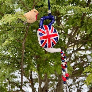 May include: A white ceramic teapot ornament with a Union Jack design hangs from a tree branch. The teapot has a red, white, and blue beaded chain hanging from the spout. A small brown bird ornament is perched on the branch.