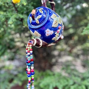 May include: A blue ceramic teapot bird feeder with floral and berry designs, suspended from a tree branch. A beaded stream of colorful beads flows from the spout. A yellow bird is perched on a nearby branch.