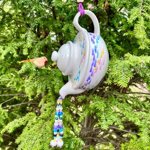 May include: A white ceramic teapot with colorful brushstroke designs, suspended by a purple cord and wooden bead. A beaded tassel flows from the spout. A small wooden bird is perched nearby, set against a backdrop of green foliage.