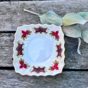 May include: A square white porcelain dish with scalloped edges and a gold-trimmed rim. The dish features a repeating pattern of red maple leaves and black and red plaid rectangular motifs with gold scrollwork accents on the interior surface.