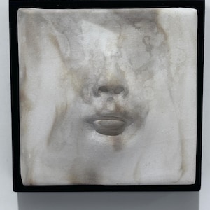 May include: A square wall sculpture featuring a white, marble-like relief of a human nose and mouth centered on a flat surface. The relief has subtle brown and grey veining throughout the material. The entire piece is framed in a thin black border.