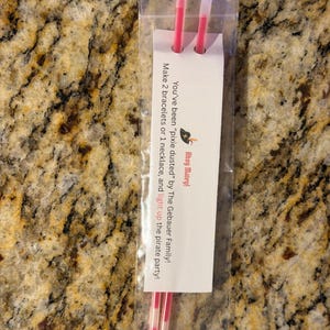 May include: Two pink glow sticks inside a clear plastic bag attached to a white card with the text: You've been "pixie dusted" by The Gebauer Family! Make 2 bracelets or 1 necklace, and light up the pirate party! Ahoy Matey!