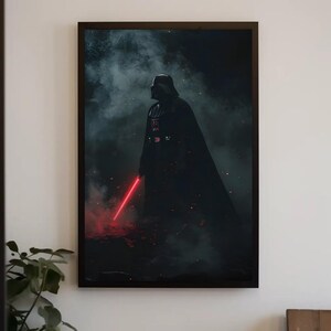 Op de afbeelding: Een ingelijste rechthoekige wandkunstprint met een donkere, sfeervolle afbeelding van Darth Vader die in een rokerige, schaduwrijke omgeving staat. Het figuur draagt een zwart harnas en houdt een gloeiend rood lichtzwaard vast met zwevende as.