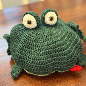 May include: A handmade, green crocheted frog with large, white-rimmed eyes and a red tongue. The frog has a textured surface and is sitting on a wooden surface. The frog is a whimsical, decorative item.