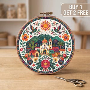 May include: A circular embroidery hoop containing a cross-stitch pattern of a Mexican-style village with a church, mountains, cacti, birds, and flowers in vibrant colours. The text BUY 1 GET 2 FREE is visible in the top right corner of the image.