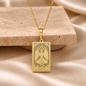 Puede incluir: Un collar con colgante rectangular de tono dorado que presenta un diseño grabado de la carta del tarot El Diablo. El colgante muestra una figura alada con cuernos y una llama en la cabeza, con el número romano XV y el texto THE DEVIL.