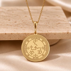May include: A gold-toned circular pendant necklace featuring an engraved sigil at the center. The perimeter of the disc displays the text SAMAEL, JACH, ELEH, and ADONAY, separated by small cross symbols. The pendant hangs from a delicate gold chain.