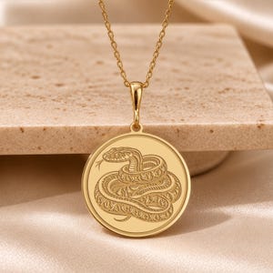 May include: A gold-toned circular pendant necklace featuring a detailed engraved design of a coiled snake with its tongue extended. The pendant hangs from a delicate matching gold-toned chain, showcasing a polished finish and intricate line work.