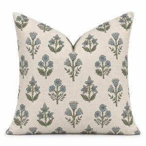 Peut inclure: Un coussin carré avec un fond beige clair présentant un motif répétitif de fleurs bleues et de feuilles vertes. Le design floral se compose de fleurs stylisées sur des tiges avec un petit feuillage, disposées en grille symétrique sur le tissu.