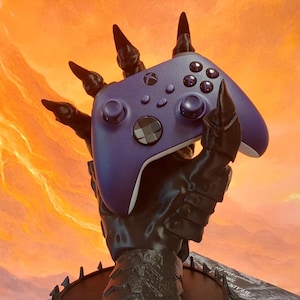 May include: A purple wireless video game controller held by a black, clawed, armoured gauntlet stand. The gauntlet features sharp, pointed finger tips and a circular base with spikes. The controller has a central Xbox logo and standard buttons.