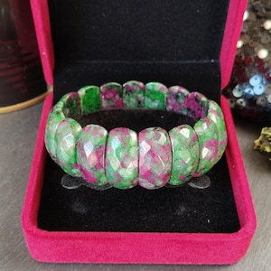 Pode incluir: Uma pulseira elástica composta por contas retangulares facetadas com um padrão manchado de rosa magenta vibrante e verde floresta profundo. A pulseira é exibida dentro de uma caixa de joias retangular de veludo vermelho brilhante.
