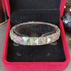 Pode incluir: Uma pulseira composta por pedras de zoisite facetadas de corte quadrado, apresentando um padrão manchado de magenta profundo e verde floresta. As pedras estão ligadas numa banda circular contínua, exibindo um acabamento polido e geométrico.