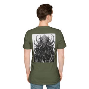 May include: An olive green short-sleeve t-shirt featuring a large rectangular black and white graphic on the back. The print depicts a detailed, tentacled creature looming over a small silhouette of a person standing in a dark, cavernous space.