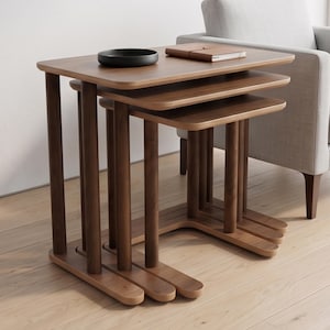 May include: A set of three nesting tables crafted from dark brown wood, featuring rectangular tops with rounded corners and cylindrical legs. The tables are stacked together, with a small black circular bowl and a brown notebook on top.