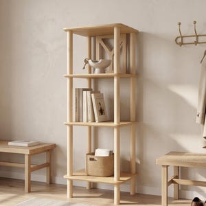 May include: A tall, light-colored wooden shelving unit with four tiers supported by cylindrical corner posts. The shelves hold a decorative bowl, several books, and a woven basket. The structure features rounded corners and a smooth finish.