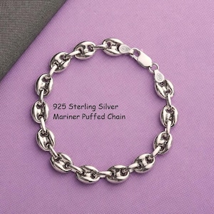 May include: A 925 sterling silver mariner puffed chain bracelet with polished oval links. The metal has a bright, reflective finish. The text 925 Sterling Silver Mariner Puffed Chain is printed in black near the center of the circular chain.