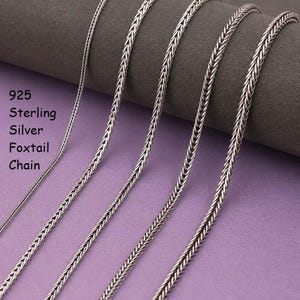May include: Five silver foxtail chains of varying thicknesses arranged vertically. The text 925 Sterling Silver Foxtail Chain is printed on the left side. Each chain features a woven, braided metal link pattern with a polished, metallic finish.