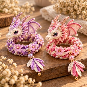 May include: Two crocheted dragon bracelets, one in shades of purple and one in shades of pink. Each features a dragon head with large purple eyes, small wings, and a coiled body decorated with crocheted flowers, white pearls, and a dangling leaf charm.